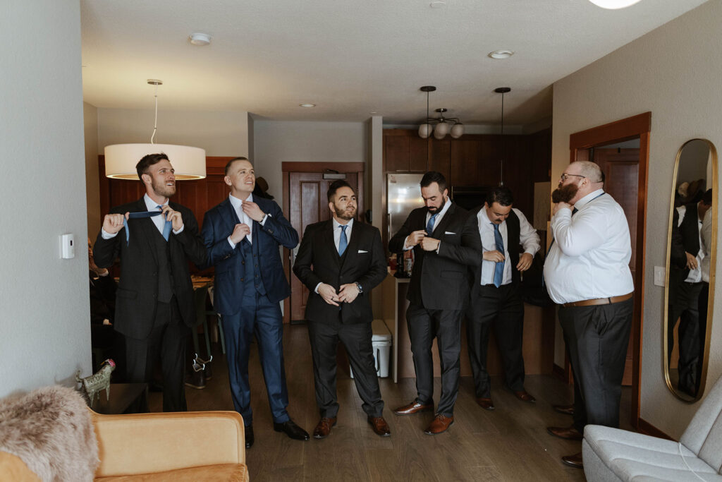 Groom in blue suit and groomsmen getting ready in hotel room together at Palisades Tahoe with dark wood cabinetry behind them