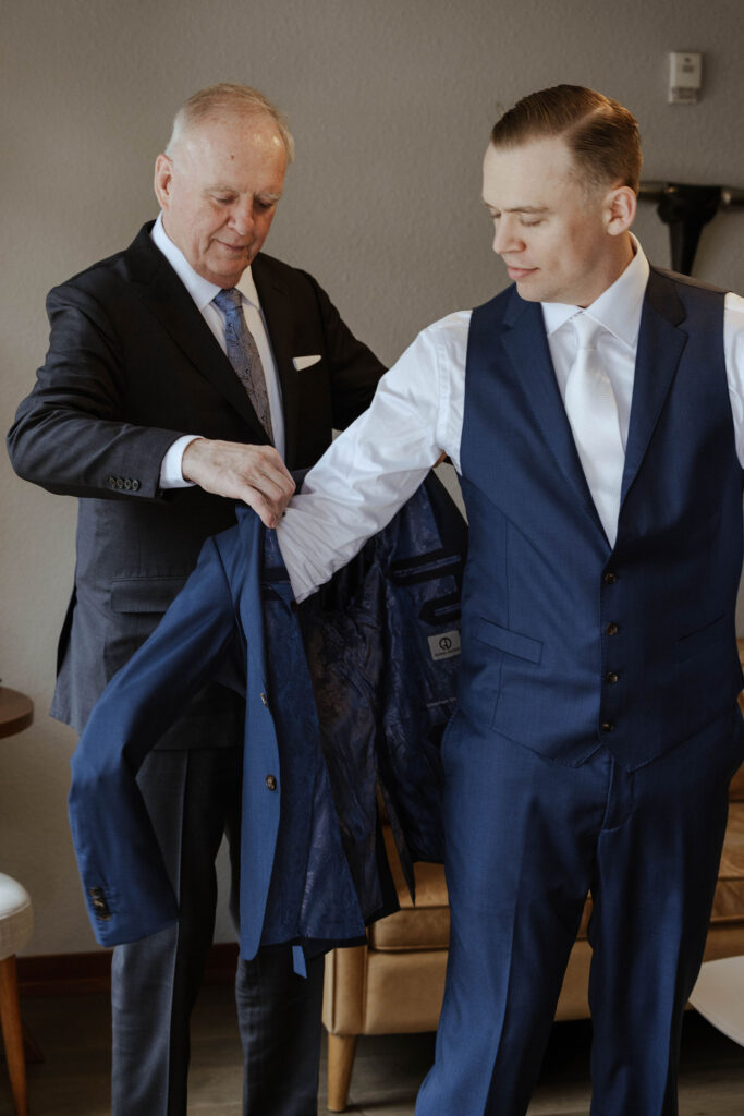 Groom's dad helping groom put on blue wedding jacket in hotel room at Palisades Tahoe