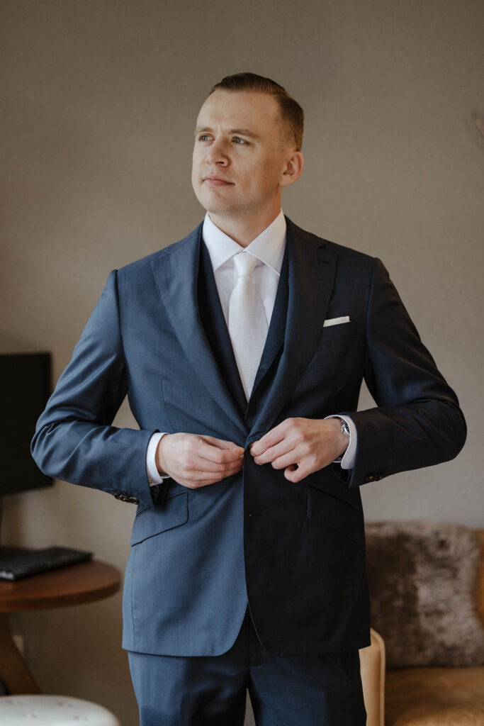 Groom in blue suit buttoning jacket while looking to side inside hotel room at Palisades Tahoe