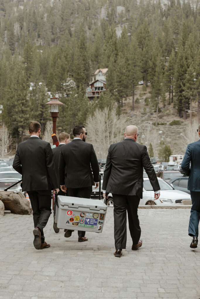 Groomsmen carrying cooler covered in stickers together across brick walkway at Palisades Tahoe