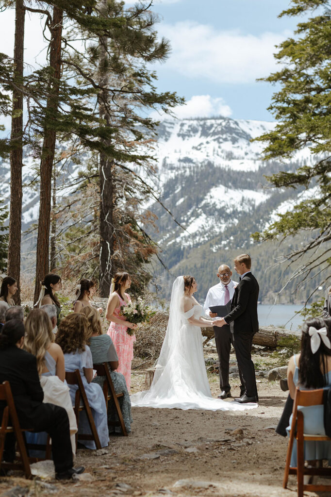 Wedding couple holding hands during ceremony in South Lake Tahoe with tall pine trees and snowy mountains in background