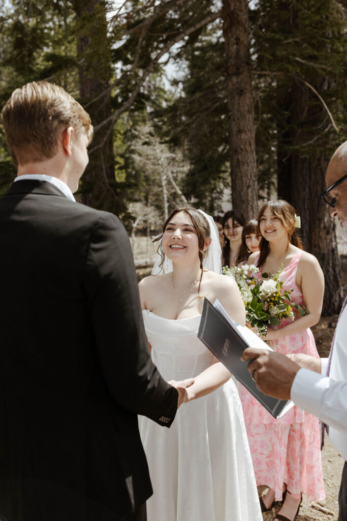 Bride smiling while holding hands with groom during wedding ceremony in South Lake Tahoe with bridesmaids and pine trees in background