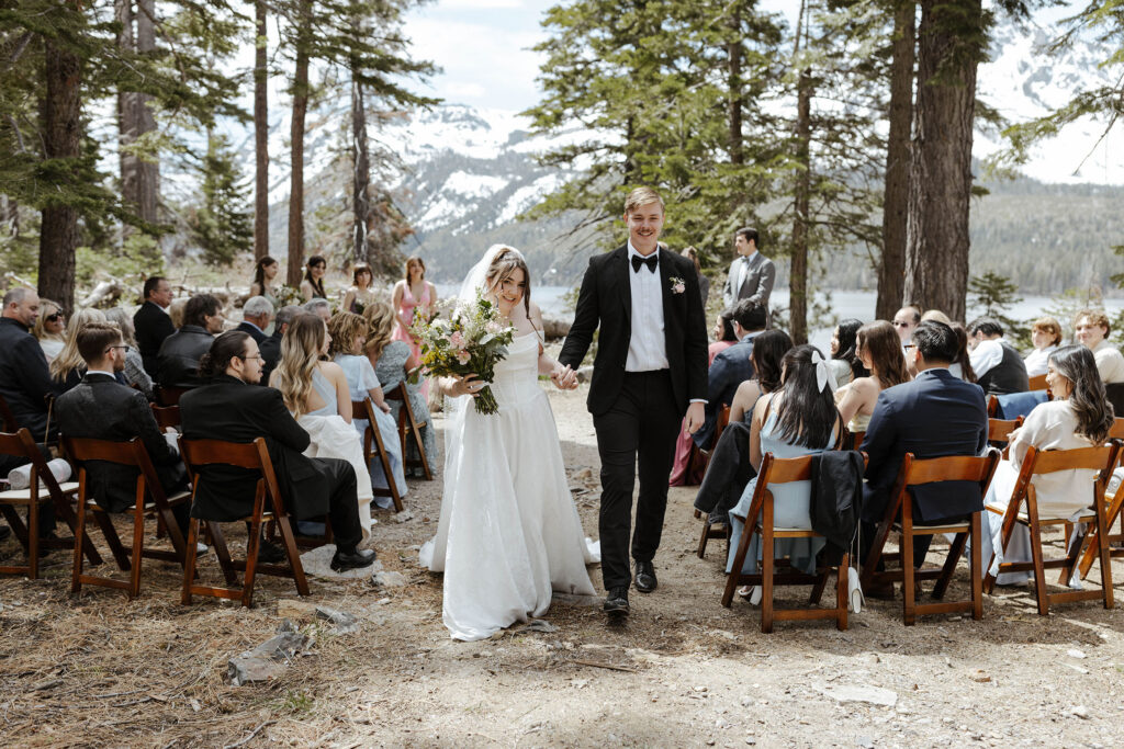Wedding couple holding hands while smiling and walking out of ceremony together in South Lake Tahoe with tall pine trees and snowy mountain in background