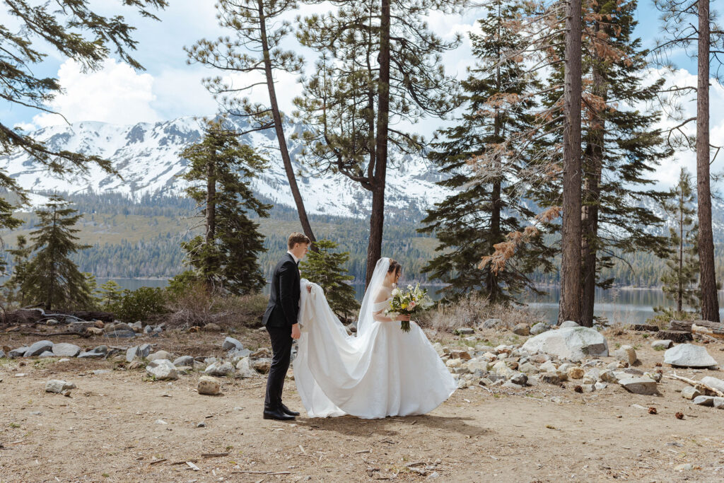 Groom holding up brides wedding dress as they walk across dirt trail in front of pine trees in South Lake Tahoe with snowy mountains and forest in background