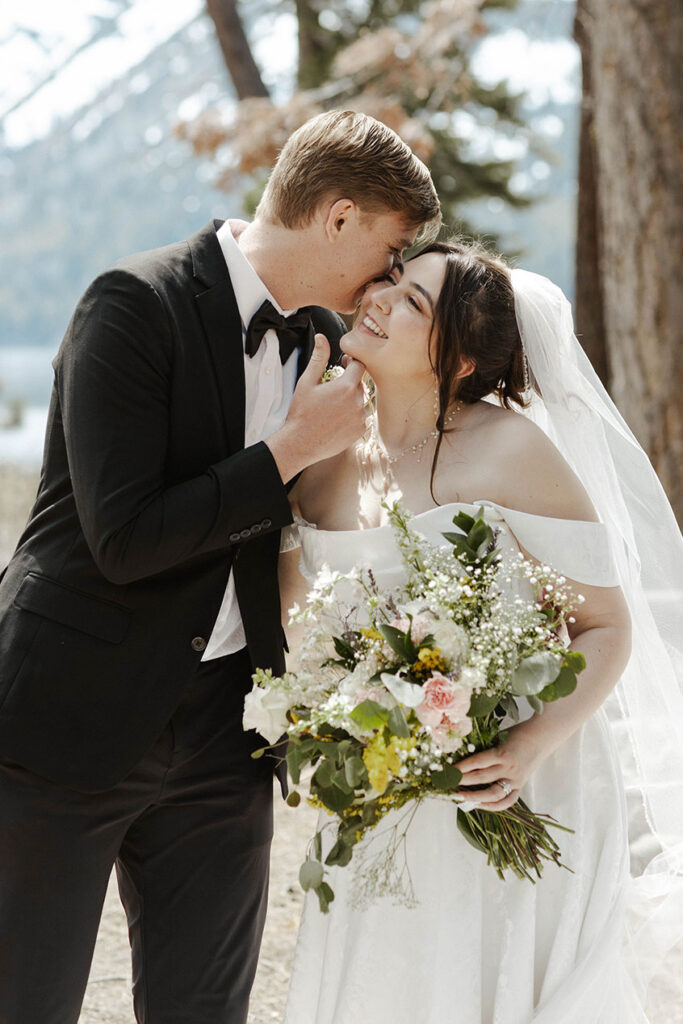 Bride smiling and holding floral bouquet as groom holds her chin and whispers in her ear in South Lake Tahoe