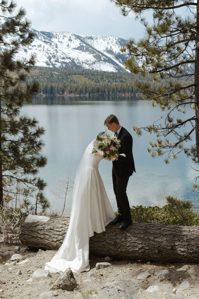 Groom helping bride stand on log while holding floral bouquet in South Lake Tahoe with lake and snowy mountains in background