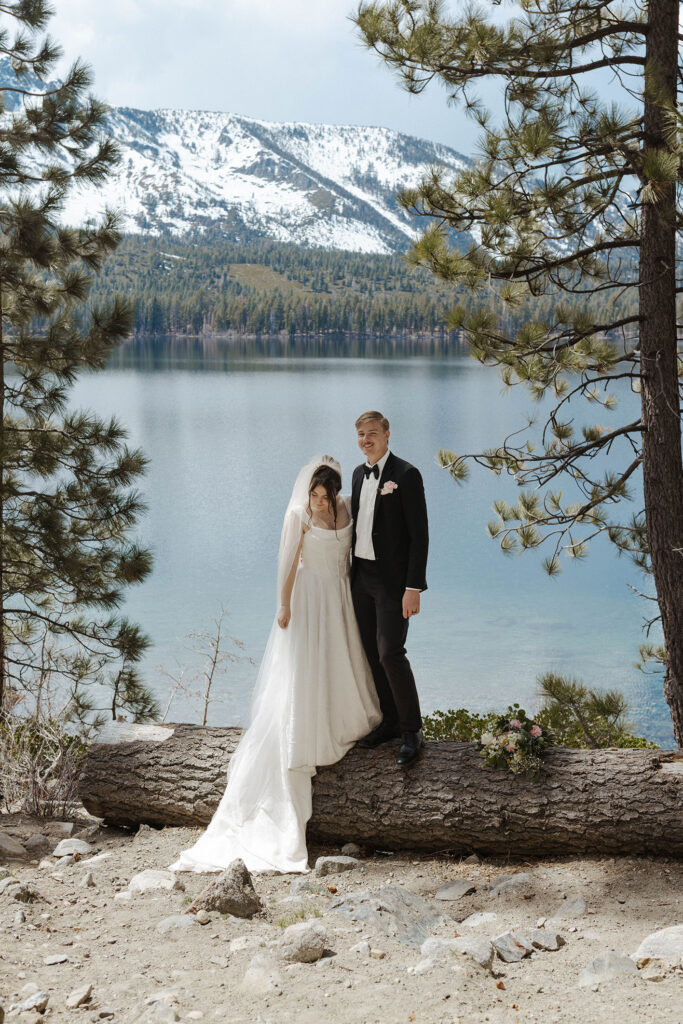 Wedding couple standing on log together in between pine trees with Lake Tahoe and snowy mountains behind them
