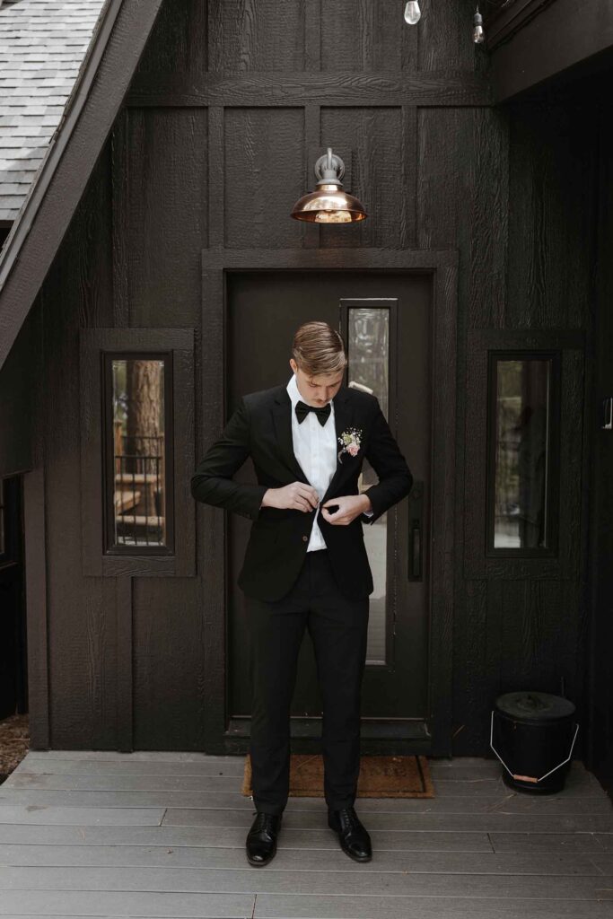 Groom standing in front of dark wooden building while buttoning his wedding jacket in South Lake Tahoe