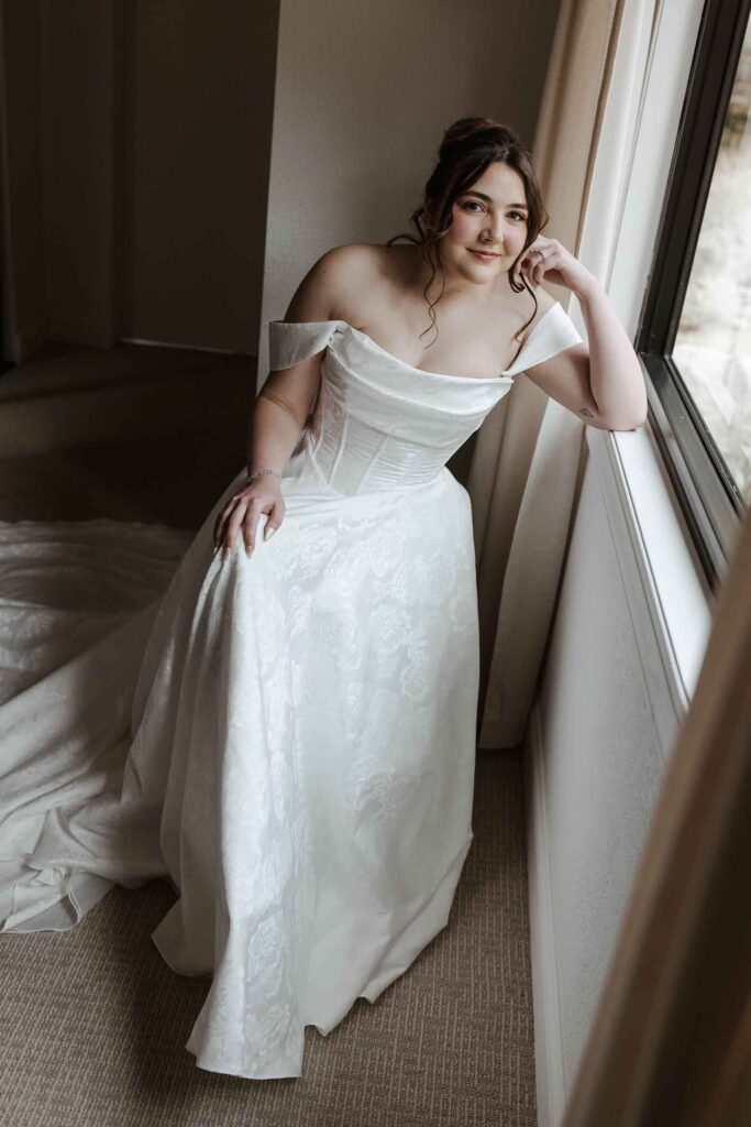 Bride in wedding dress sitting in chair and looking at camera while leaning on windowsill