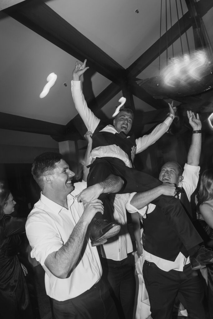 groom laughing throwing his hands up while sitting on his buddies shoulders during a wedding reception at riva grill