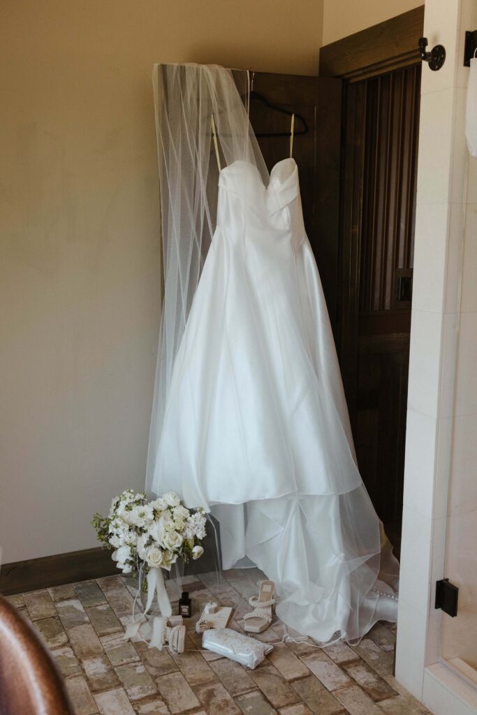 white strapless wedding dress hanging on a door with a veil and wedding details 