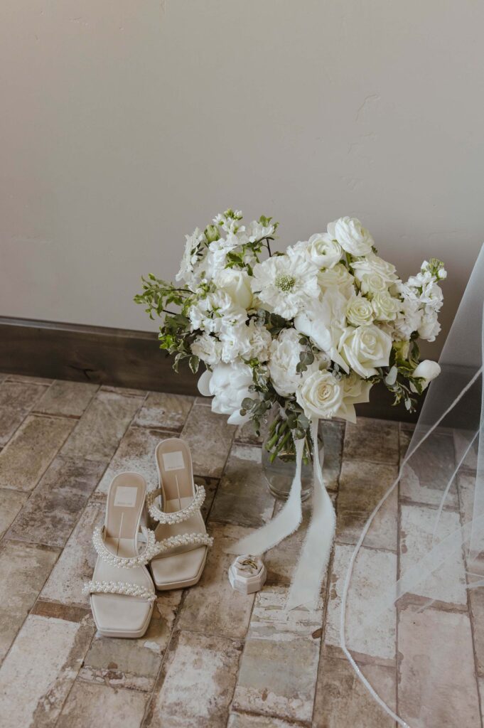 wedding bouquet with white roses and greenery, with beaded wedding heels and wedding rings laying on the tile ground