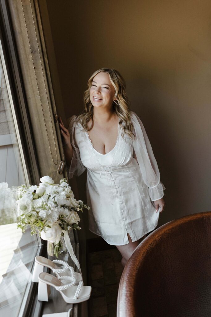 bride in white slip dress smiling and holding the window with bouquet and shoes in the window 
