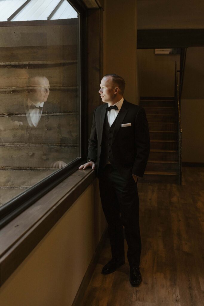 groom standing looking out the window with his hand on the banister where you can see his reflection