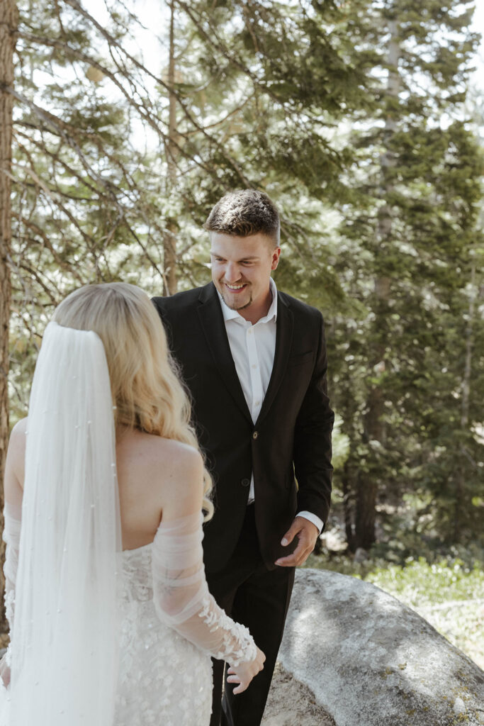 Groom smiling at bride while standing on rocks with trees in background at Emerald Bay