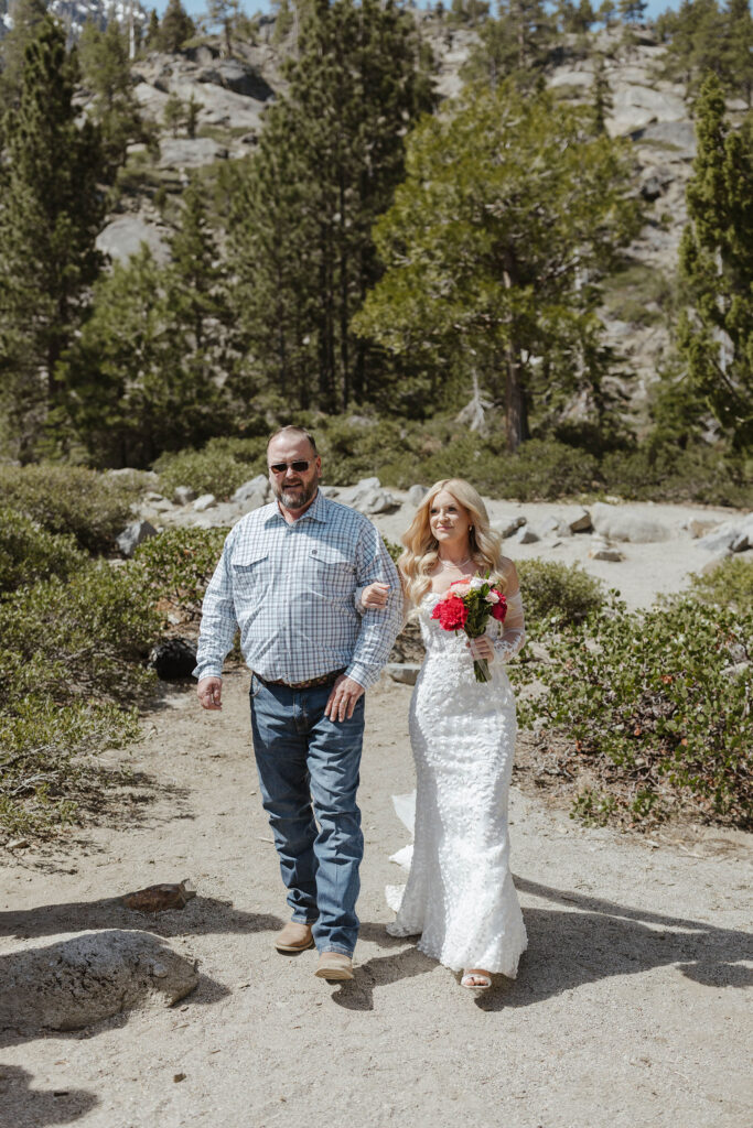 Bride holding dad's arm while walking down to elopement ceremony in Emerald Bay with trees in background