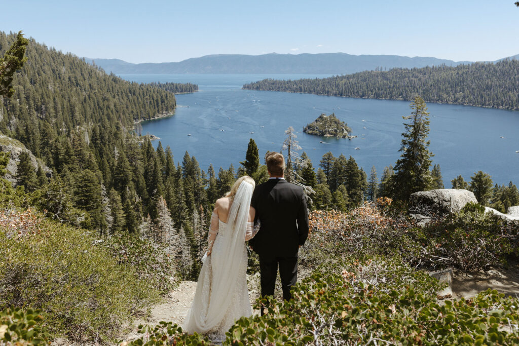 Elopement couple standing together while bride holds groom's arm and both look out over Emerald Bay at Lake Tahoe