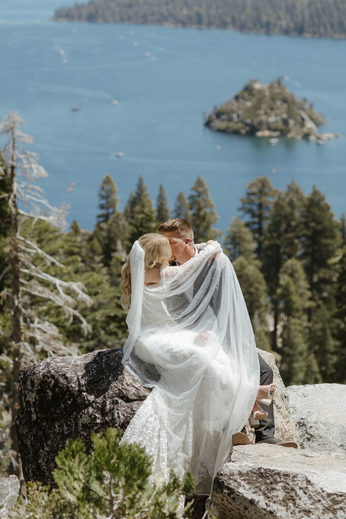 Elopement couple sitting on a rock together and kissing while bride covers groom partially with veil at Emerald Bay with Lake Tahoe in background