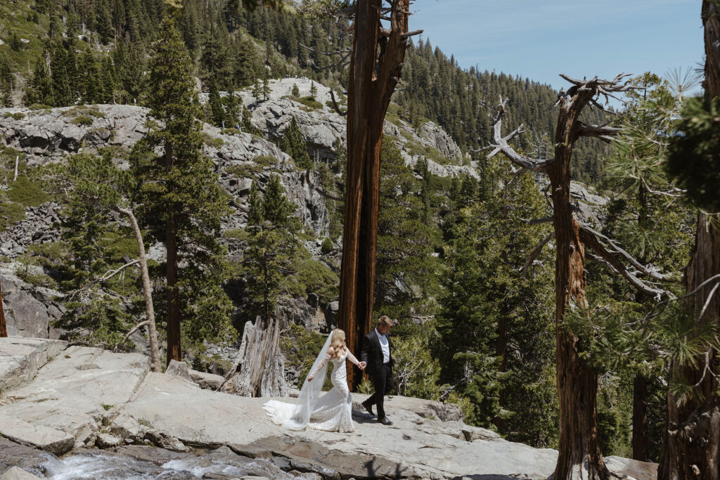 Elopement couple holding hands while walking down large rocky ledge together at Emerald Bay with trees in background