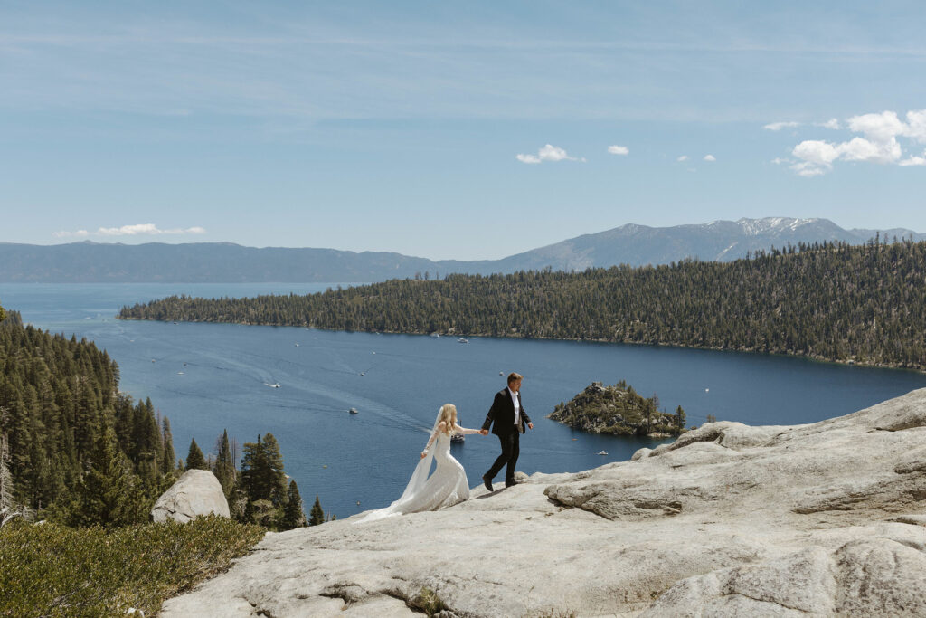 Man holding wife's hand while walking up rocks together at Emerald Bay with Lake Tahoe and pine trees in background