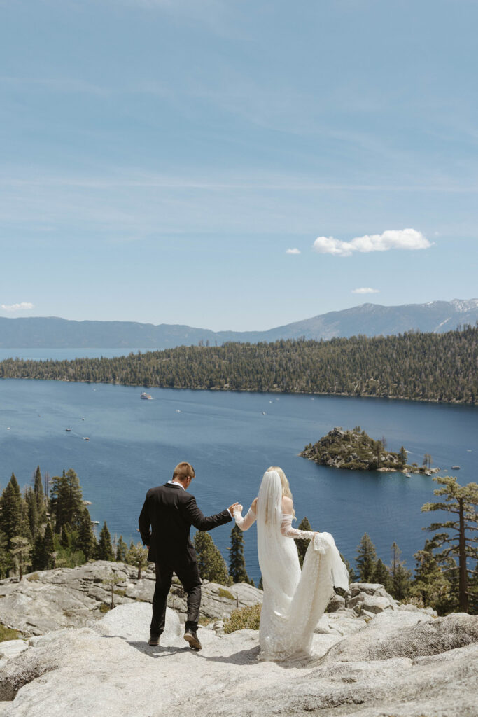 Husband holding wife's hands while walking down rocks together at Emerald Bay with Lake Tahoe in background