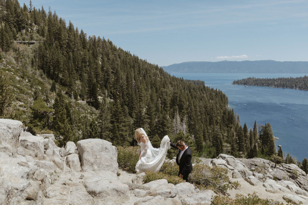 Groom holding bride's dress as both walk up rocky trail together at Emerald Bay with pine trees and Lake Tahoe in background