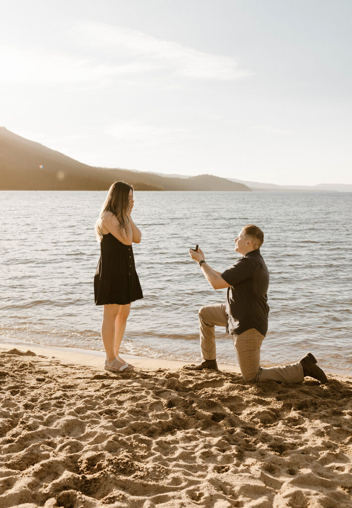 man on one knee proposing on the beach with arm extended wit ha ring proposing to woman in the summer in lake tahoe