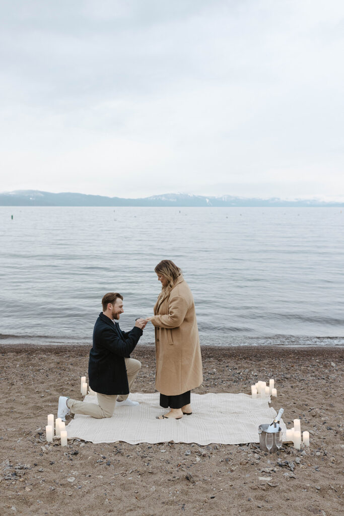 man on one knee proposing on the beach proposing to woman on a blanket with candles in lake tahoe