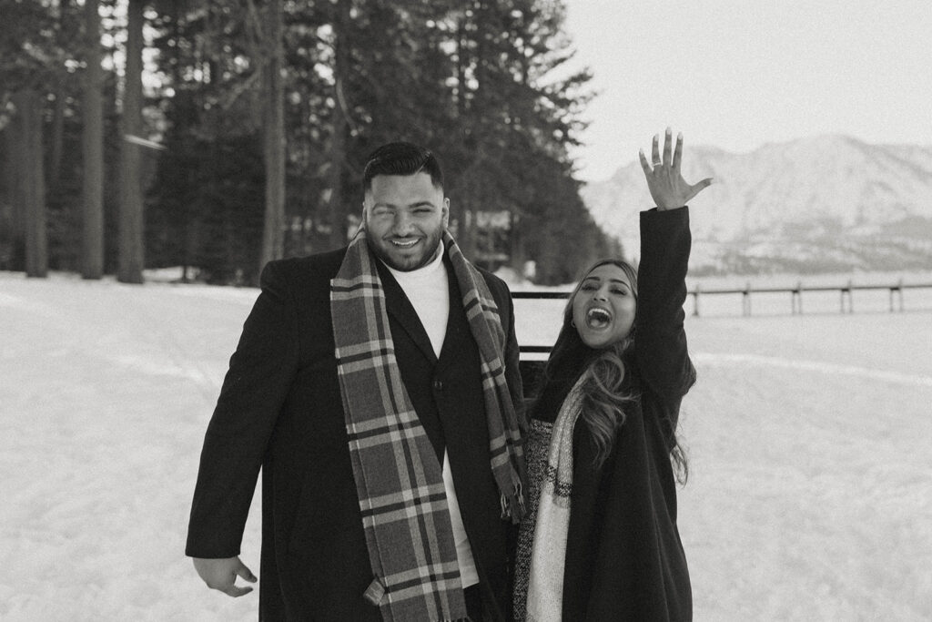 woman throwing hand up yelling she's engaged while man smiles and laughs in lake tahoe in the winter