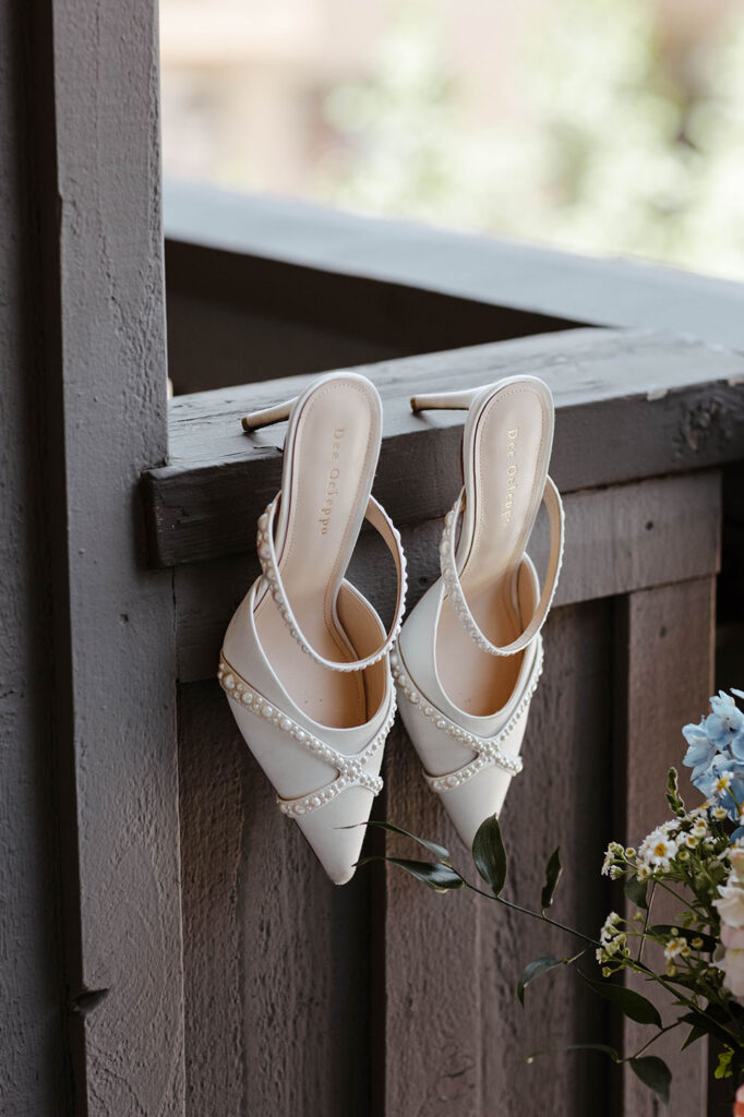 Bride's wedding shoes with pearl decorated straps hanging on side of wooden railing next to flowers at Palisades Tahoe