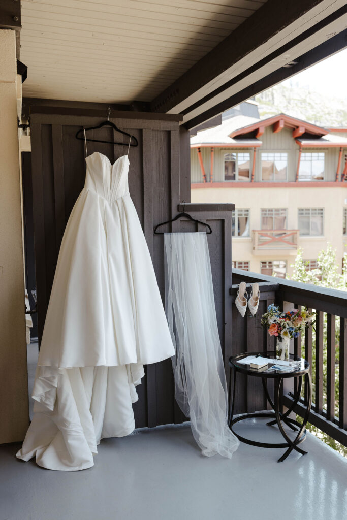Bride's wedding dress hanging on wooden wall outside next to veil and details on glass table at Palisades Tahoe
