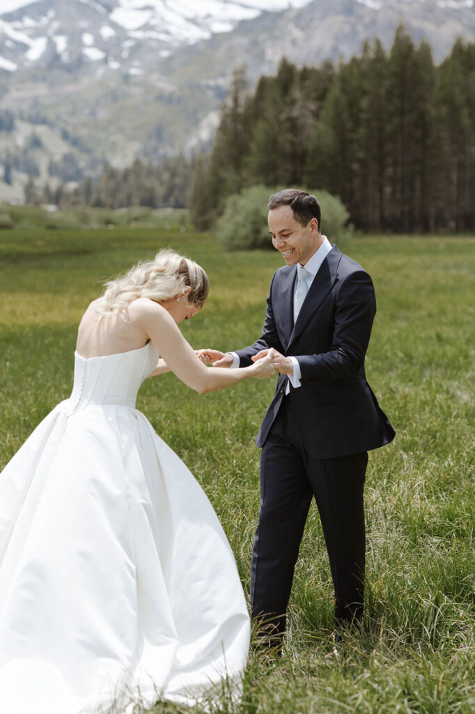 Wedding couple holding hands while standing in large grass field with tall pine trees and mountains in background at Palisades Tahoe