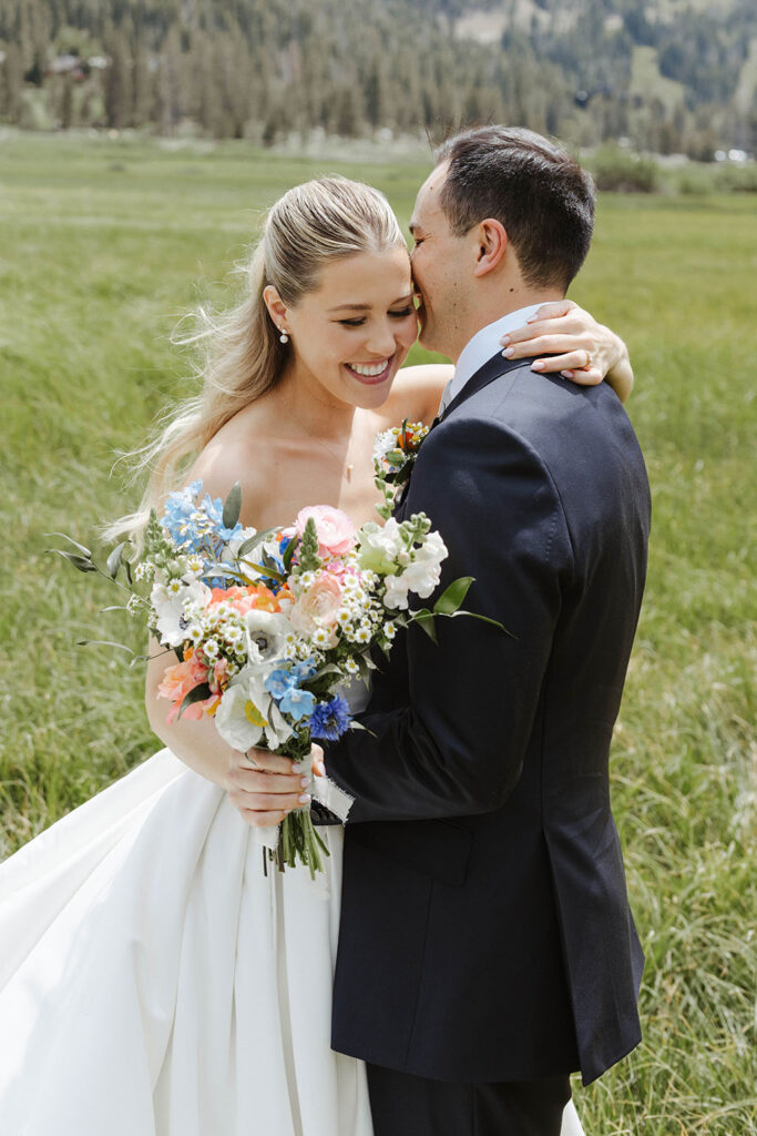 Bride smiling while being held by wedding groom and both stand in open grass field at Palisades Tahoe
