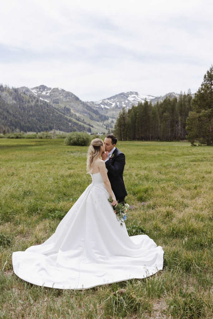 Wedding couple kissing while standing in large grass field together with tall pine trees and mountains in background at Palisades Tahoe