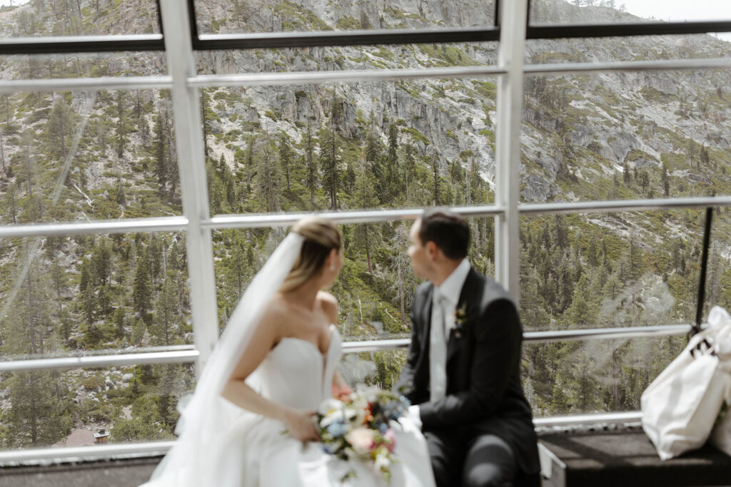 Wedding couple sitting on a bench on tram together and looking over their shoulders out window towards pine trees on mountain at Palisades Tahoe