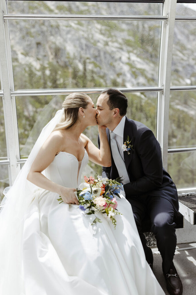 Wedding couple kissing while sitting on a bench and riding tram up Palisades Tahoe to High Camp
