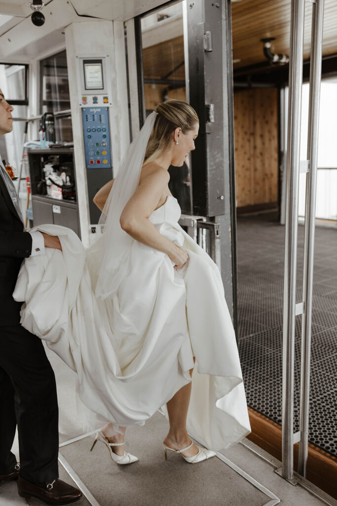 Bride holding wedding dress while stepping out of tram to High Camp