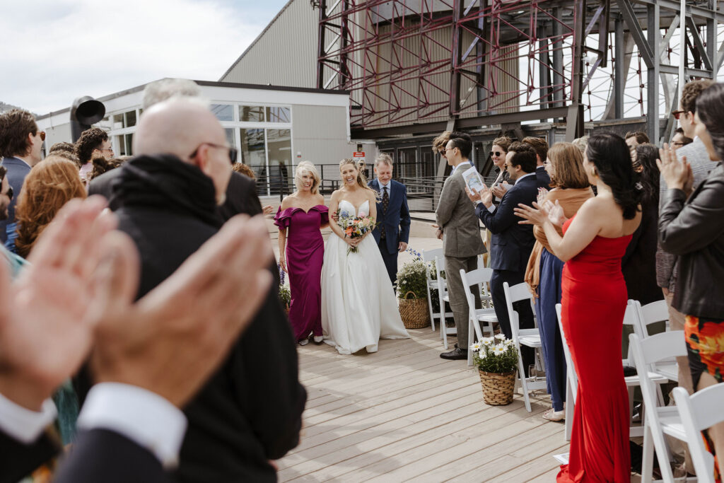 Bride holding colorful flower bouquet while walking down wedding aisle with parents to ceremony at High Camp