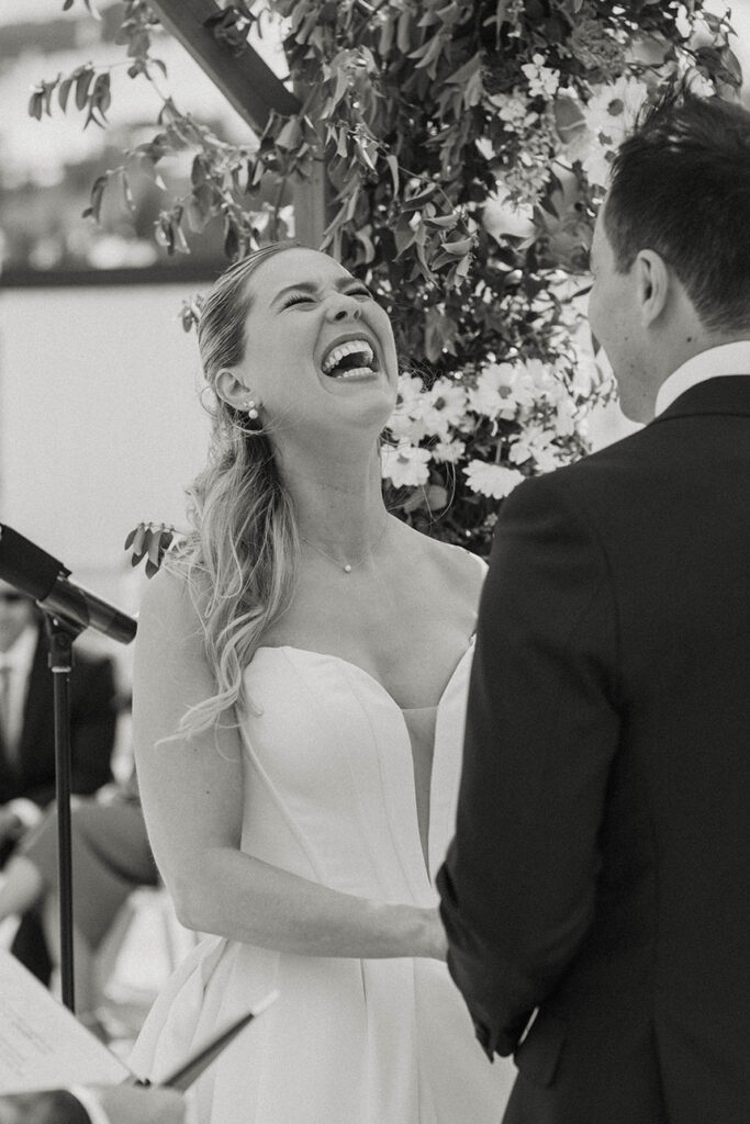 Bride laughing while holding groom's hands during wedding ceremony with floral arch in background at High Camp