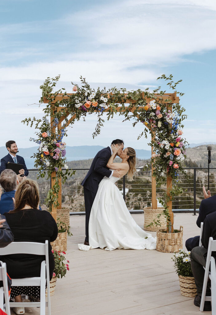 Wedding couple's first kiss after ceremony standing underneath colorful flower arch with mountains in background at High Camp