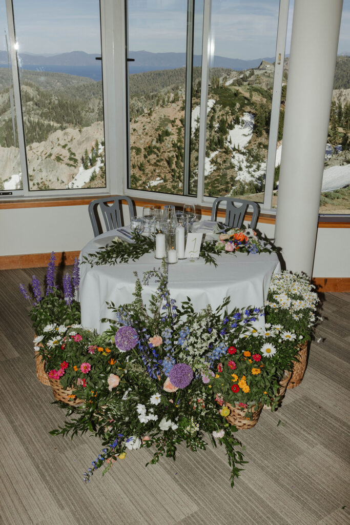 Wedding couples reception dinner table surrounded by colorful flowers with mountains visible through windows in background at Palisades Tahoe