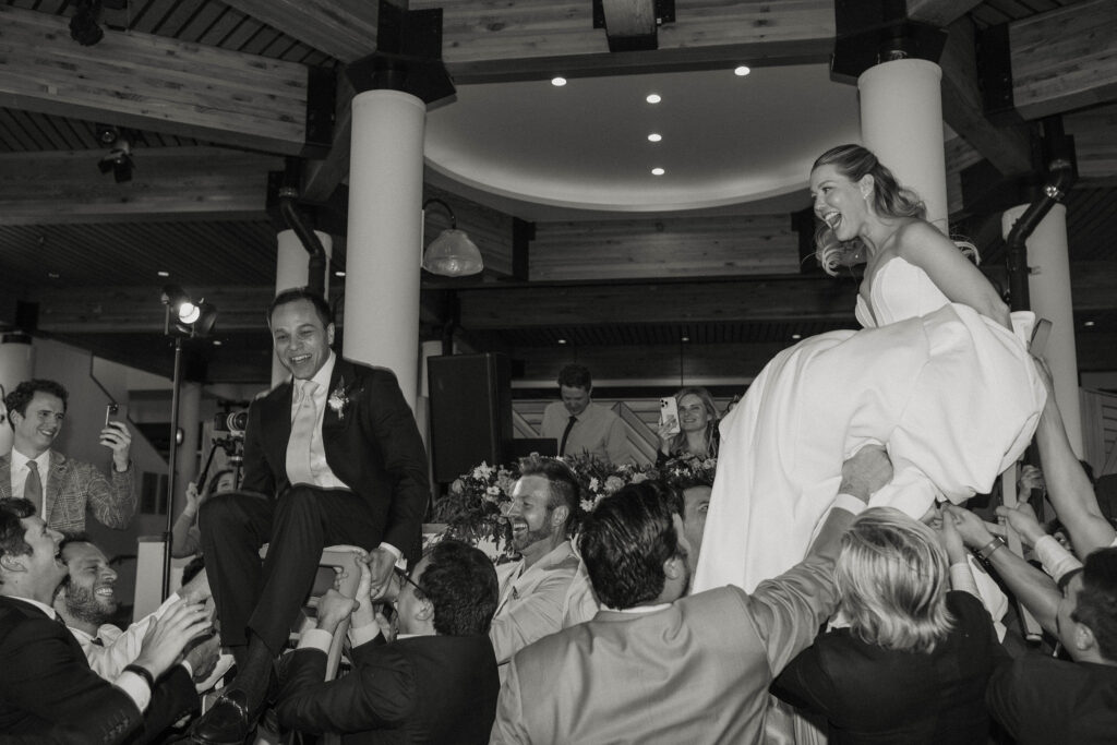 Wedding couple laughing while being lifted up on chairs by wedding guests during reception at Palisades Tahoe