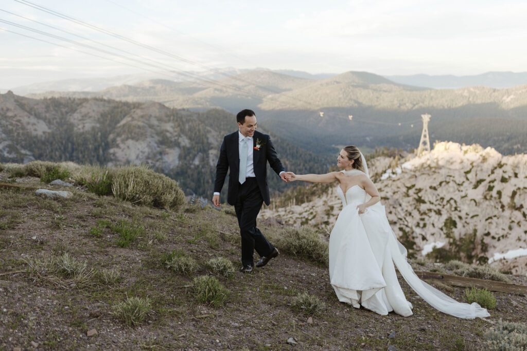 Groom holding bride's hand while smiling at her and leading her up hillside at High Camp with lots of mountains in background