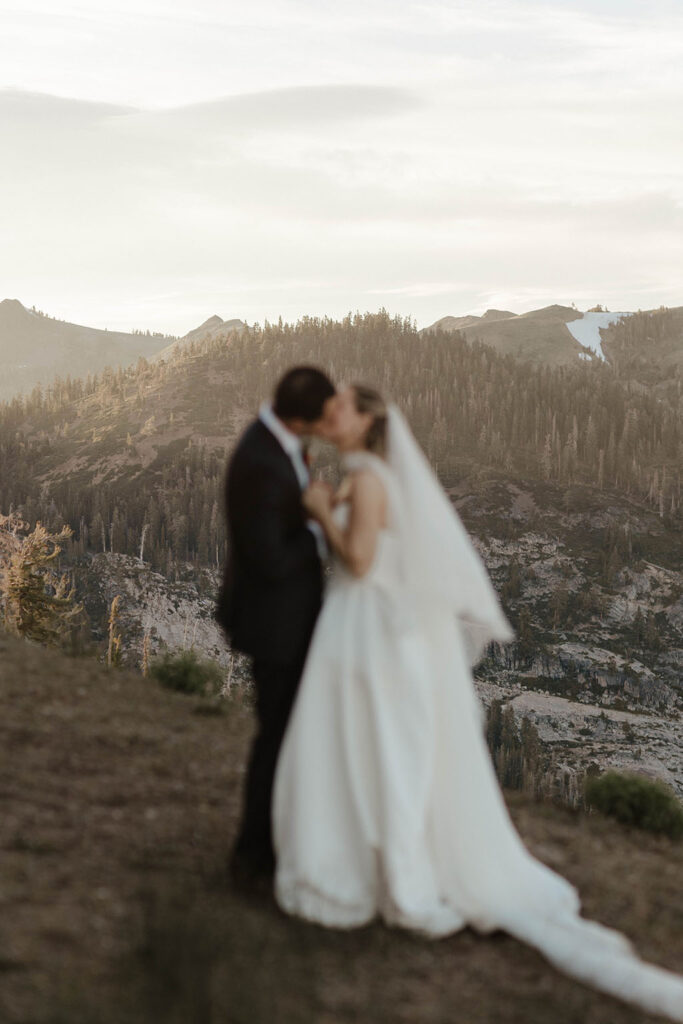 Wedding couple holding hands and kissing while standing on hillside together at High Camp with pine trees and mountains in background