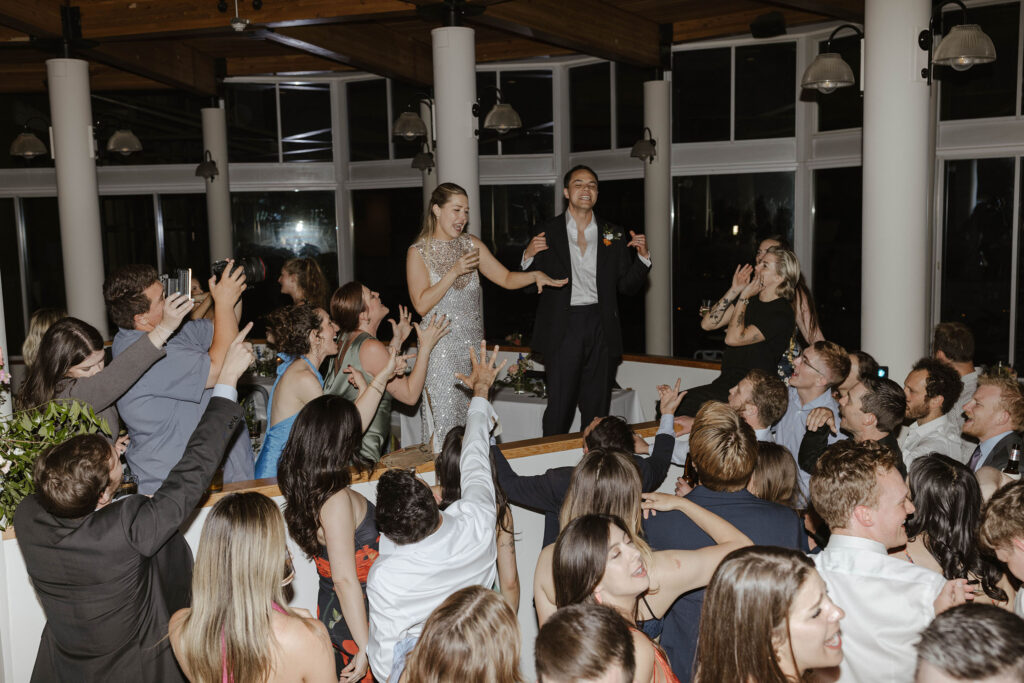 Wedding couple standing on table celebrating and singing while guests dance around them during reception at High Camp