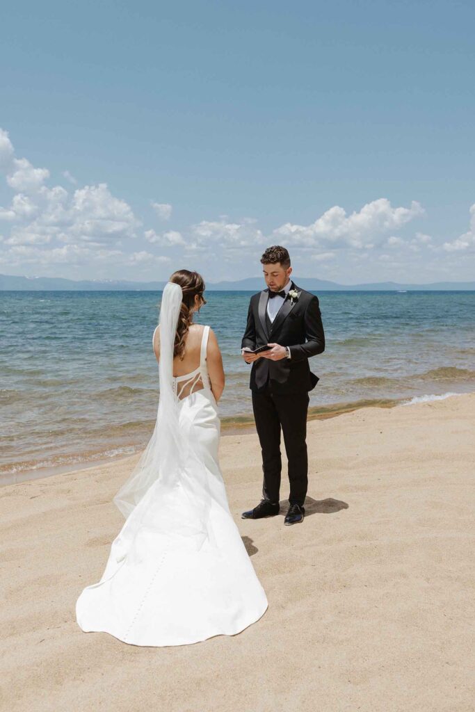 Elopement groom reading wedding vows to bride while standing on sandy beach at Emerald Bay with Lake Tahoe in background