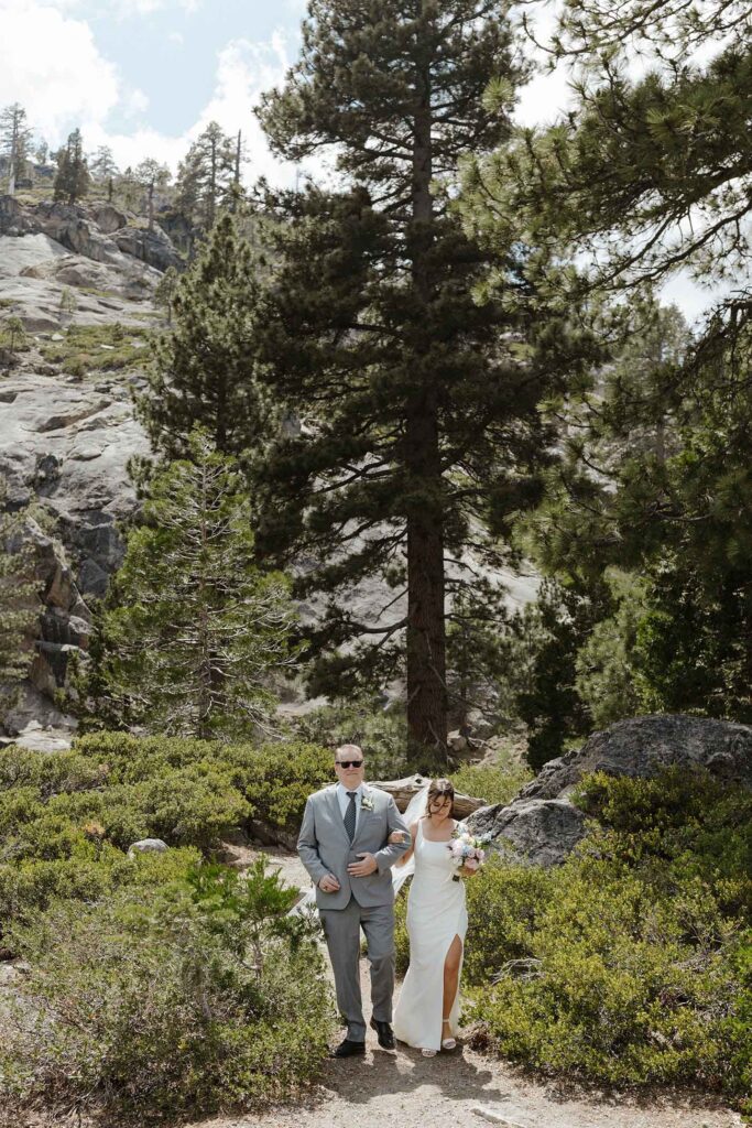 Bride holding dad's arm while walking down aisle during elopement at Emerald Bay with pine trees and mountain in background