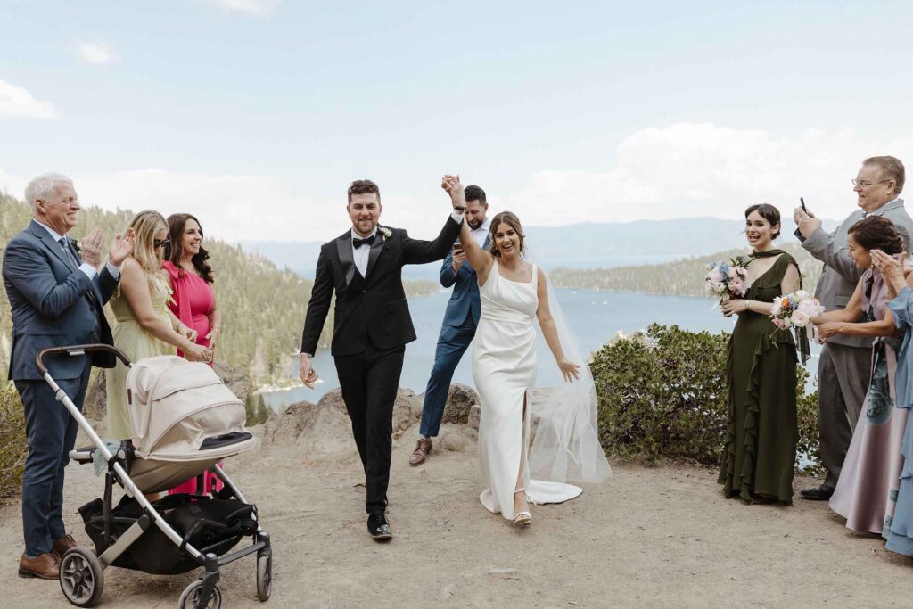 Elopement couple holding hands in the air together and smiling while walking out of ceremony at Emerald Bay while guests cheer on either side