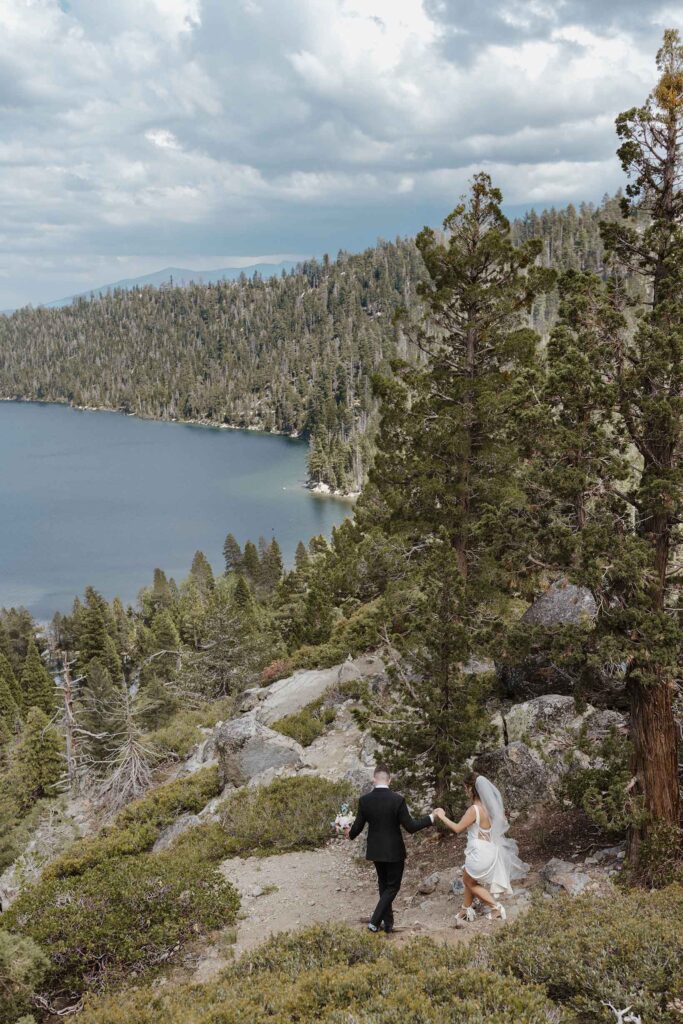 Groom holding bride's hand and helping her down trail at Emerald Bay surrounded by pine trees with Lake Tahoe in distance