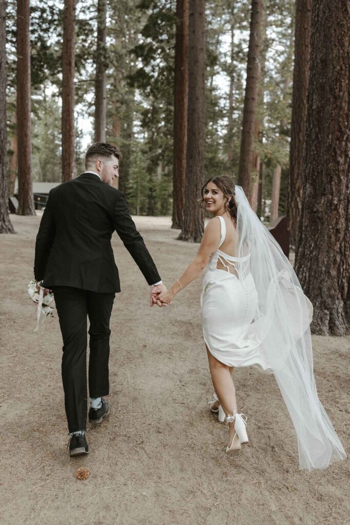 Elopement couple holding hands and walking up dirt trail together surrounded by tall trees while bride looks back over shoulder and smiles at camera at Emerald Bay